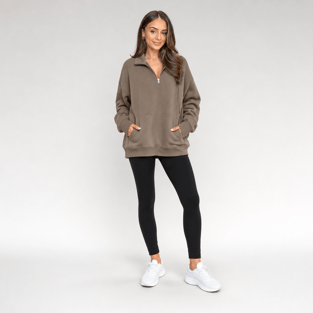 Full-body view of a female model wearing the taupe quarter-zip pullover with black trousers and white sneakers.