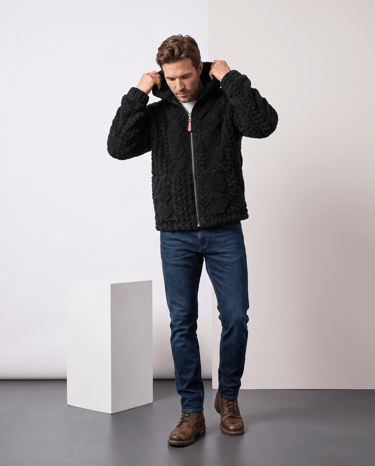 Full-length view of black hooded jacket with hands raised to the hood, jeans and brown shoes