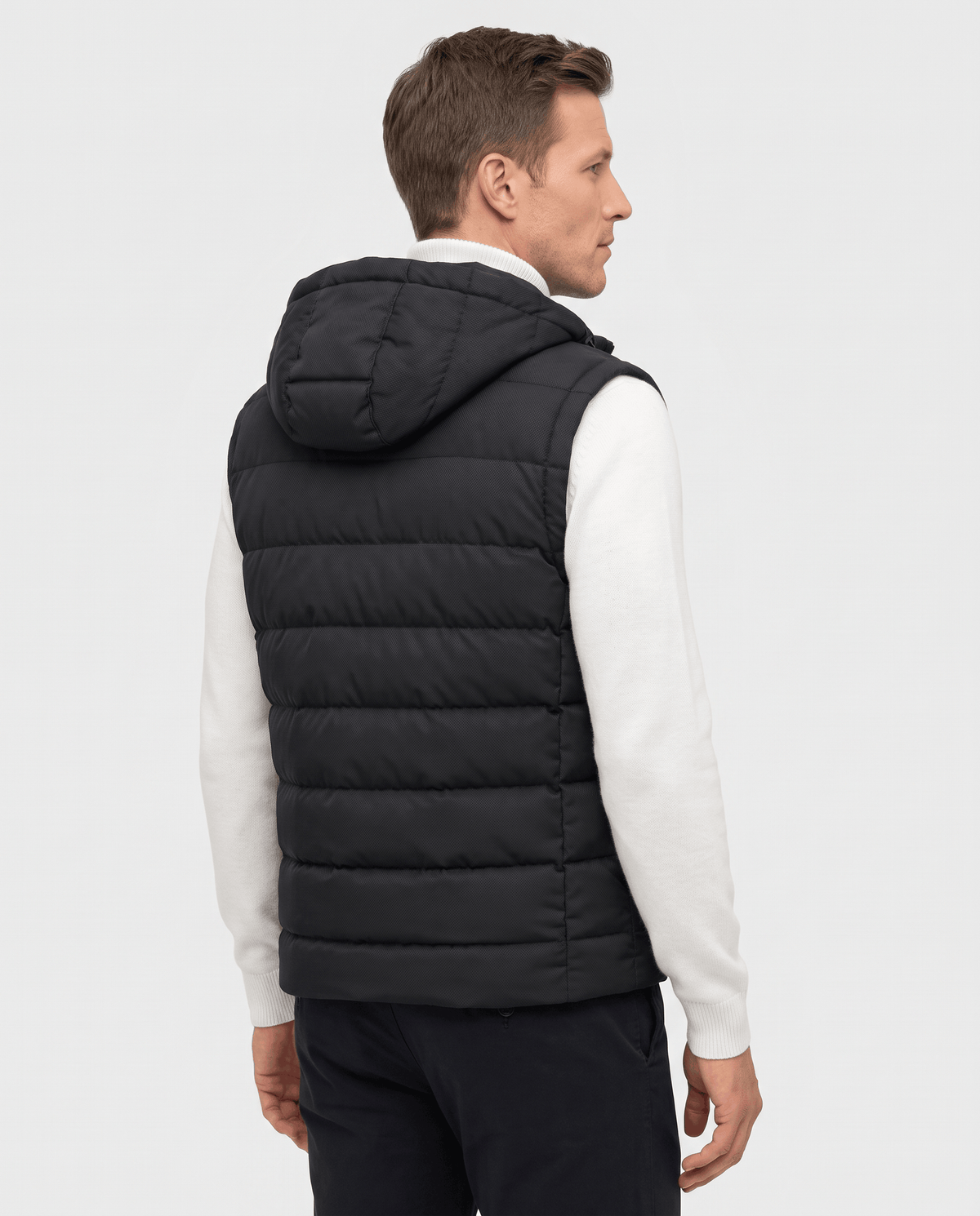 Back view of black hooded quilted vest over a white sweater and dark trousers