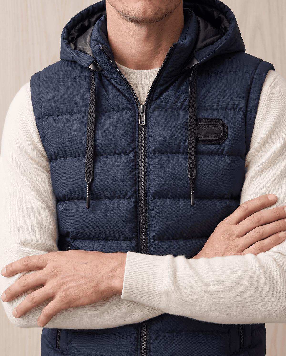 Navy quilted vest with chest badge and hood, arms crossed over a cream sweater
