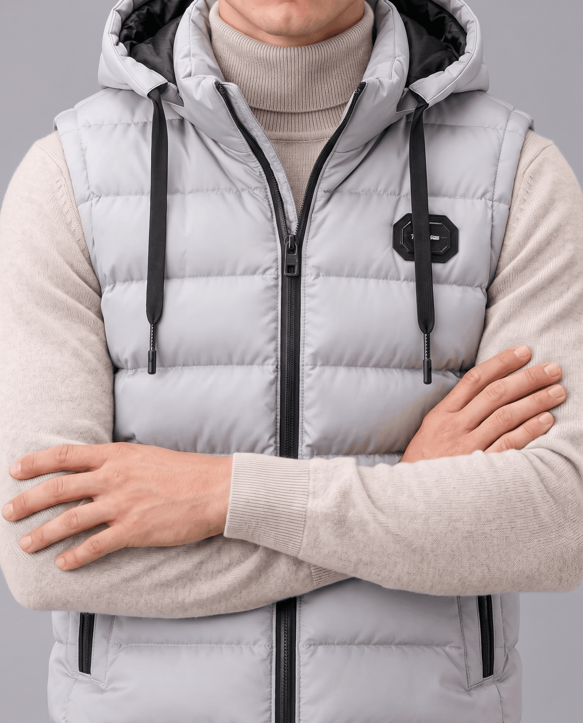 Close-up of gray vest with drawstring hood, chest badge and zip front, arms crossed