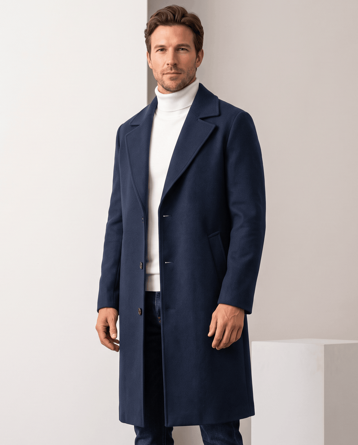 Men's longline coat in navy worn open over a white turtleneck and dark jeans, front view