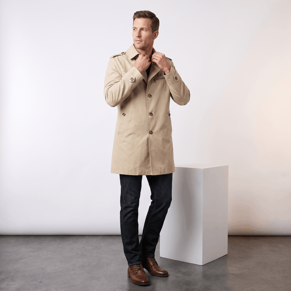 Model wearing a beige single-breasted trench coat over a dark shirt and jeans in a studio.