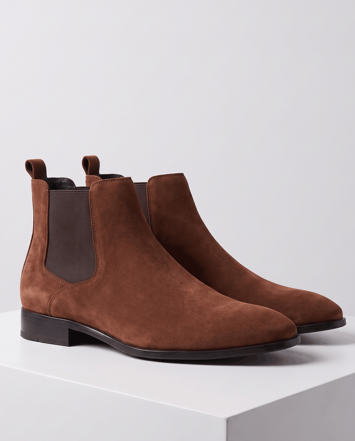 Brown suede Chelsea boots placed side by side on a light gray block