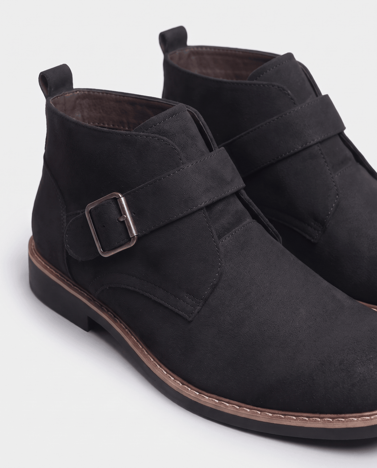 Close-up of a black suede boot with buckle strap and tan stitched sole edge