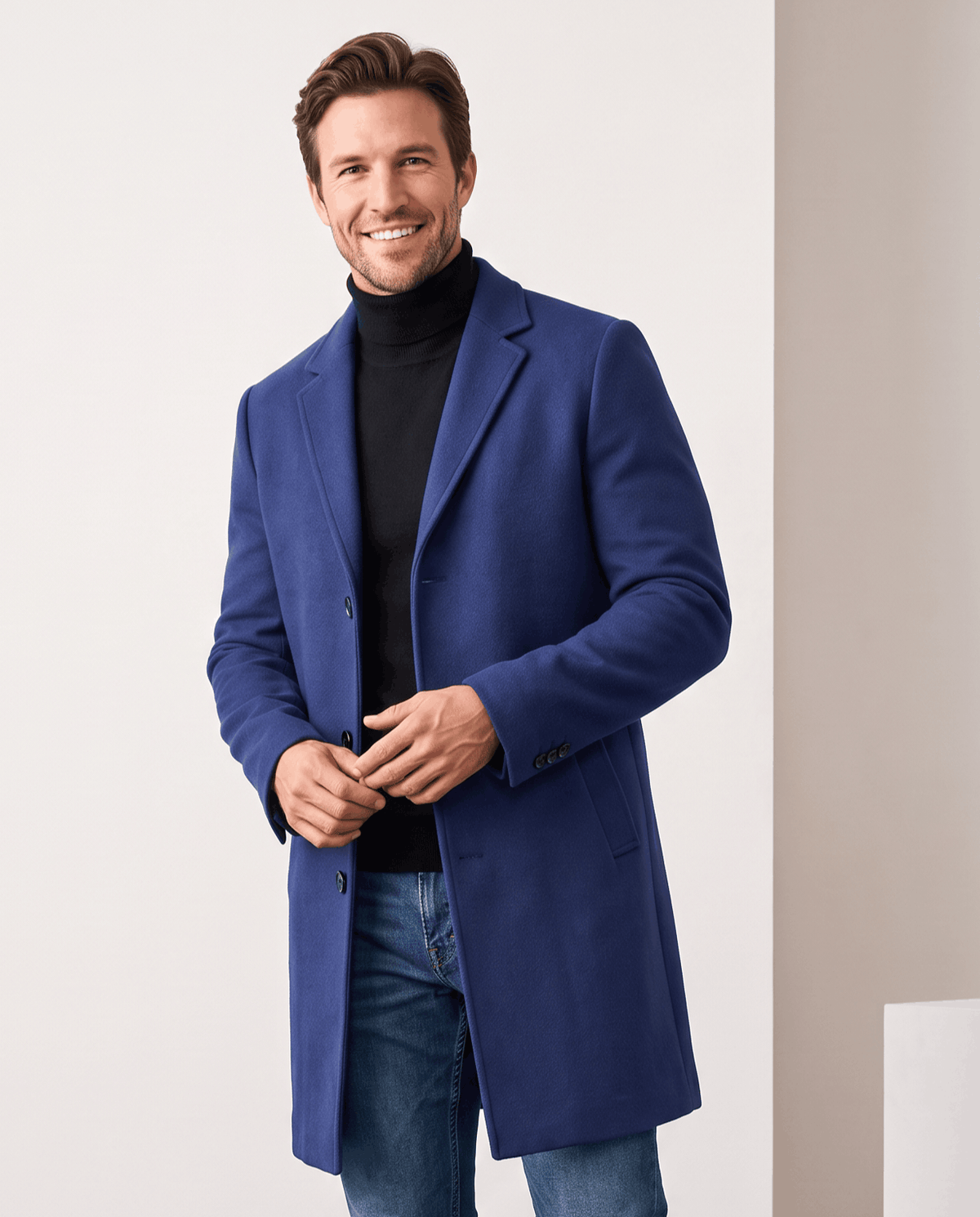 Blue tailored overcoat worn over a black turtleneck and jeans, with hands at the front