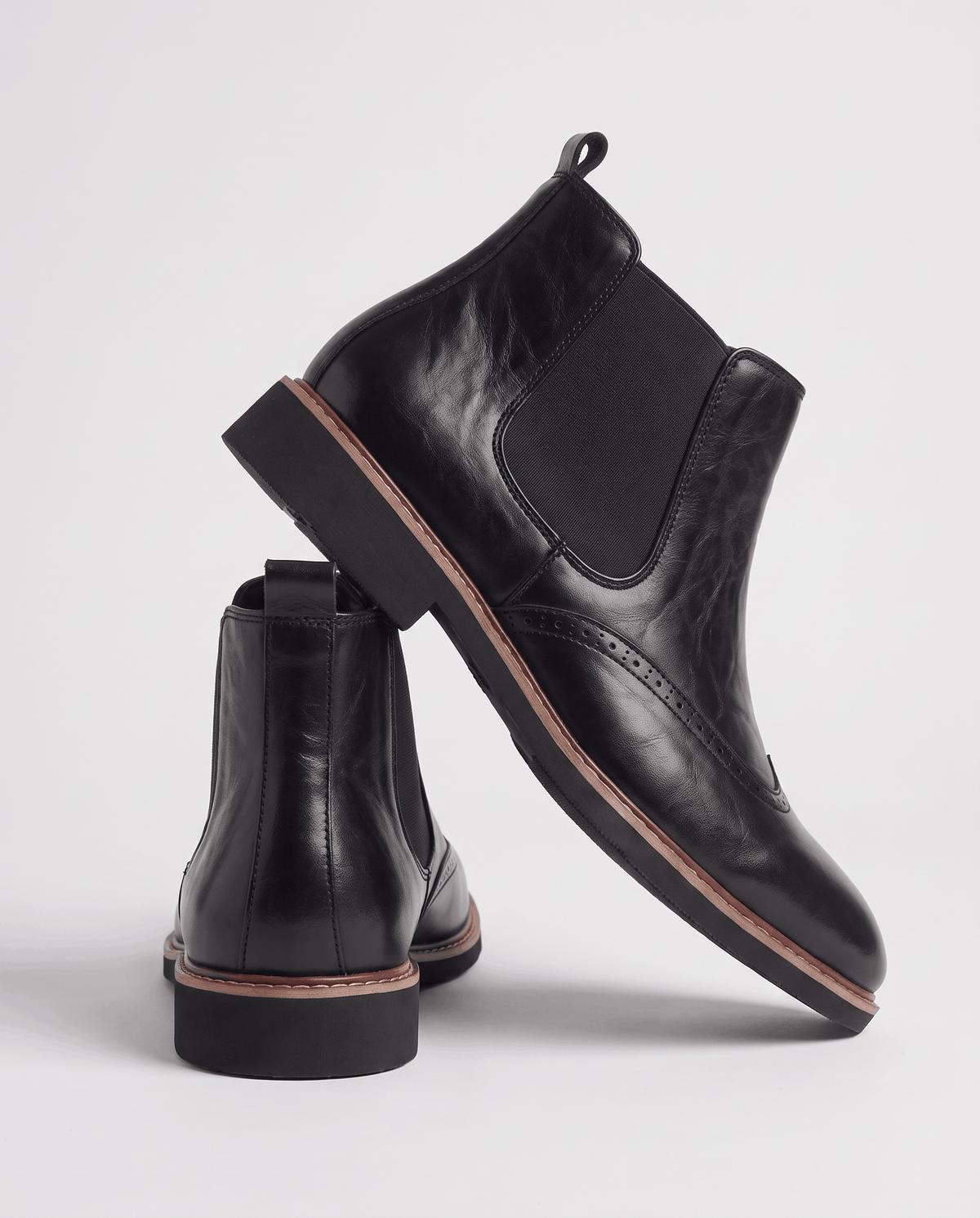 Black brogue boots stacked, one leaning on the other to show the sole and wingtip toe
