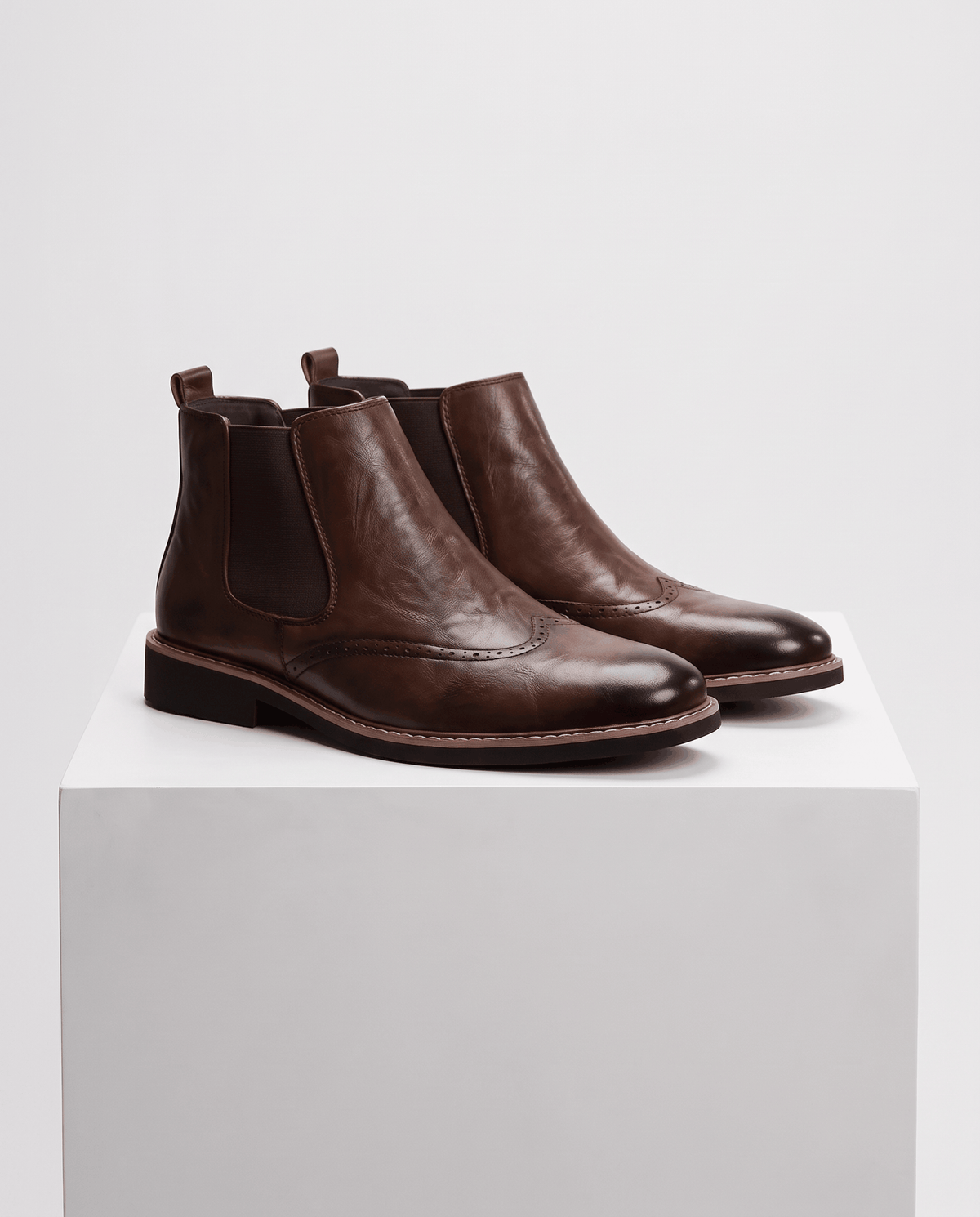 Pair of brown brogue boots with elastic side panels on a white pedestal