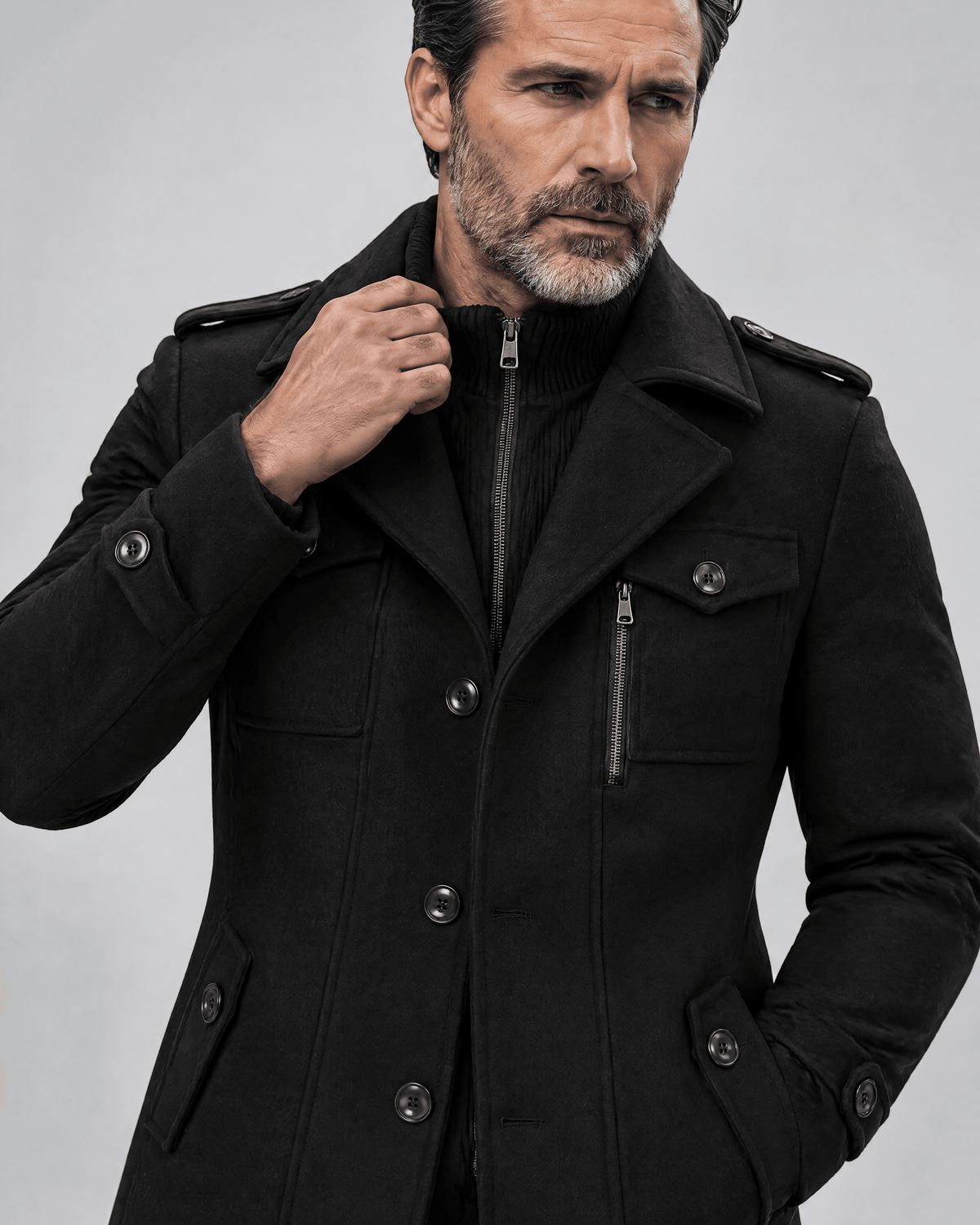 Close-up of the black coat's chest pockets, zip detail and epaulettes with a hand at the collar