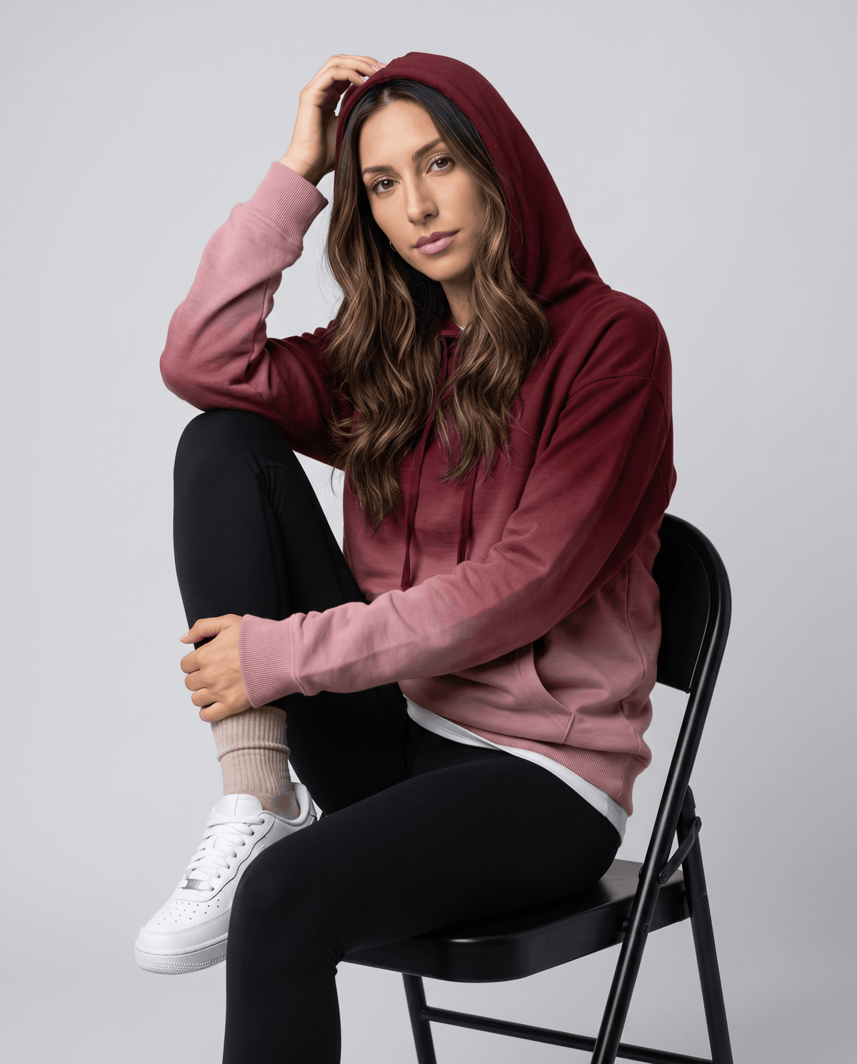 Seated look in the burgundy fade hoodie with the hood up, black leggings and white sneakers