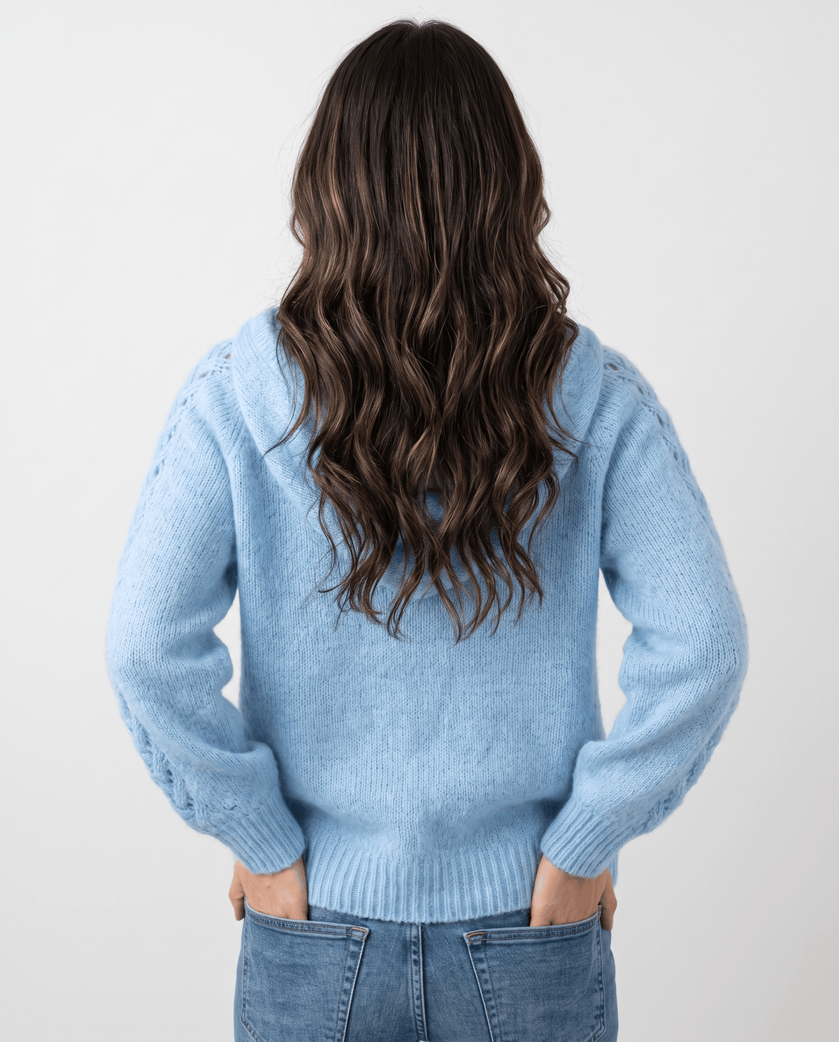 Back view of the light blue knit sweater with the hood down and hands in jeans pockets