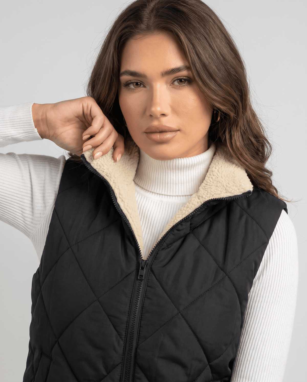 Model touching the cream collar of a black quilted vest at the zip