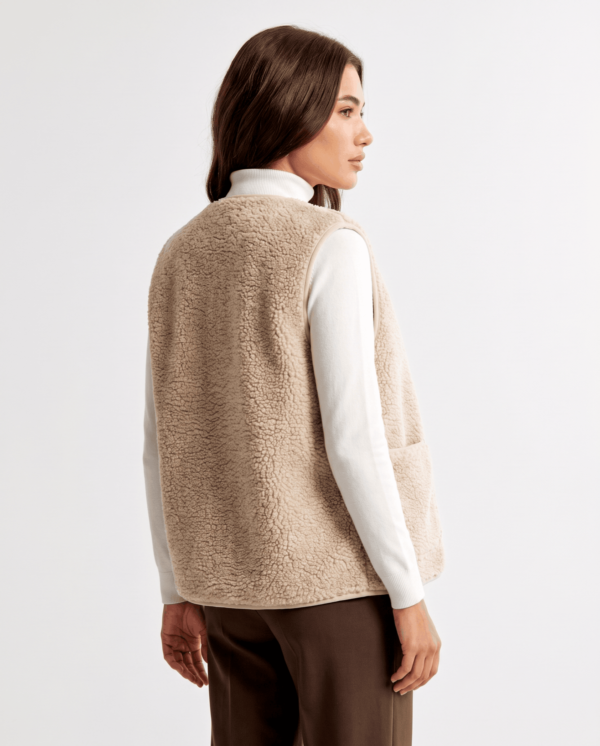 Back view of beige teddy fleece vest over a white turtleneck and brown trousers