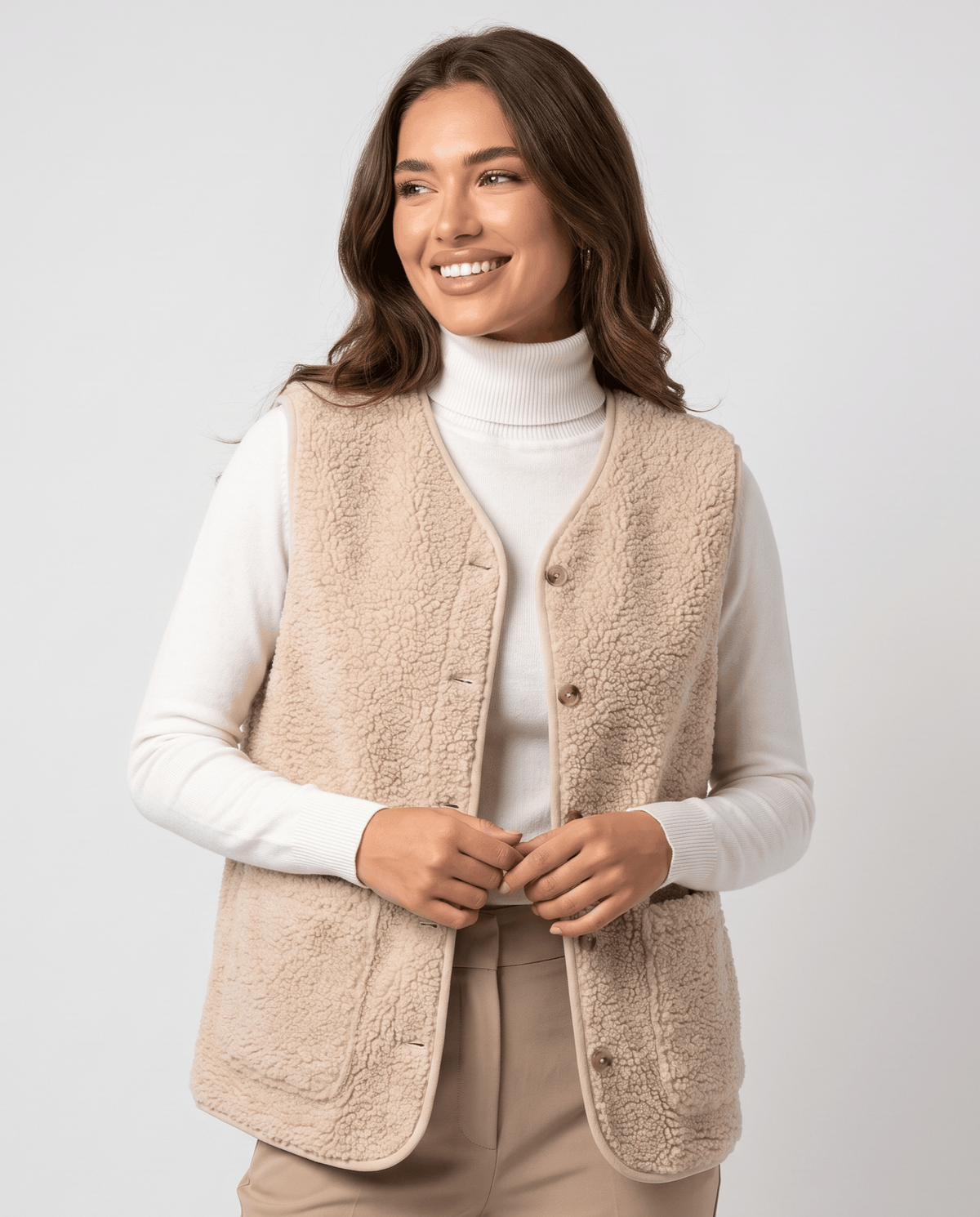 Beige teddy fleece vest buttoned at the front over a white turtleneck and tan trousers