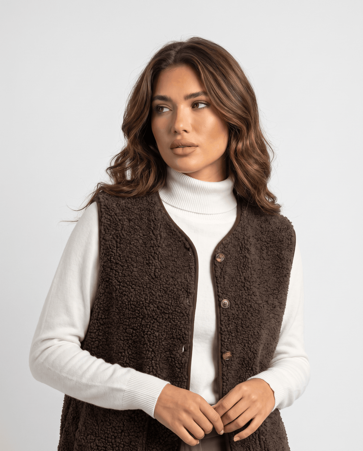 Brown teddy fleece vest with button front worn over a white turtleneck, hands at the placket