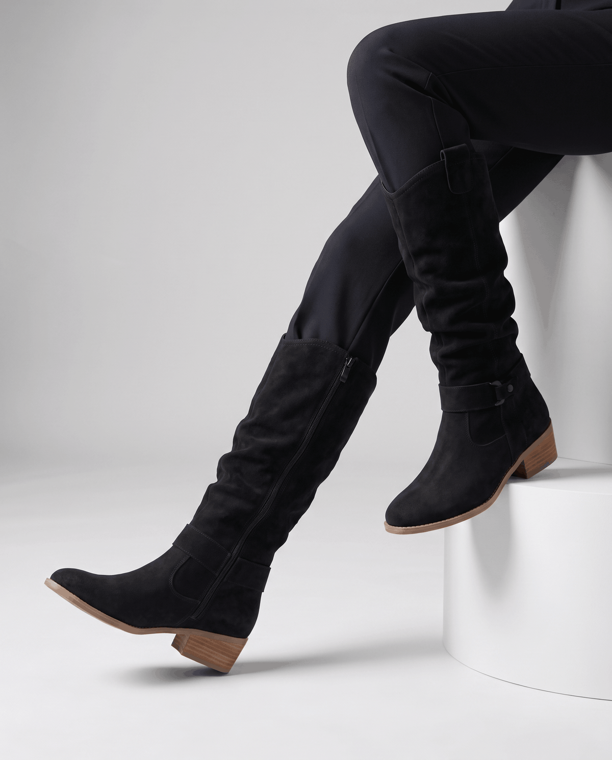 Woman seated wearing black vegan suede knee-high slouch boots with black trousers.