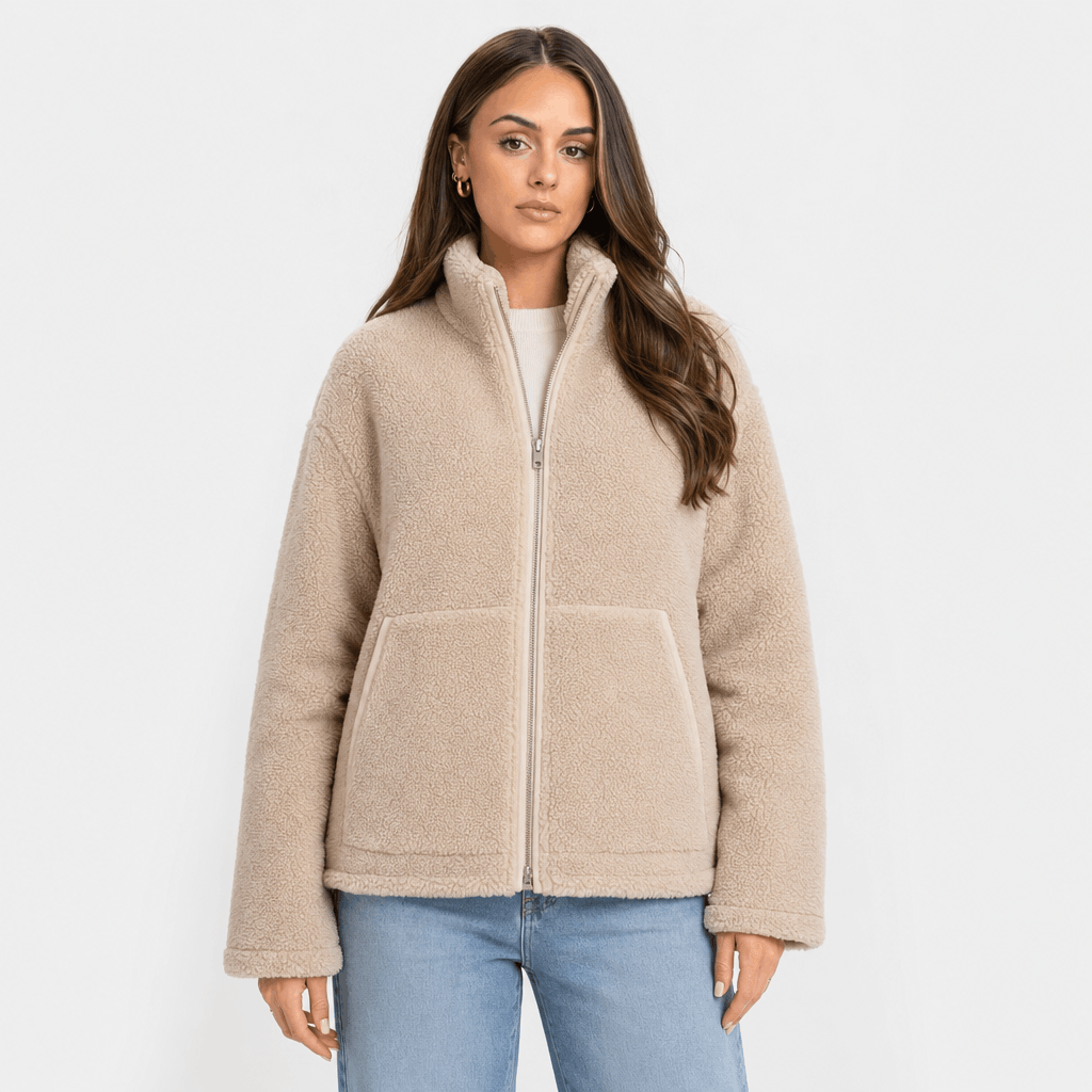 Female model wearing a light beige sherpa zip-up jacket with a high stand collar and large front patch pockets, paired with blue jeans.