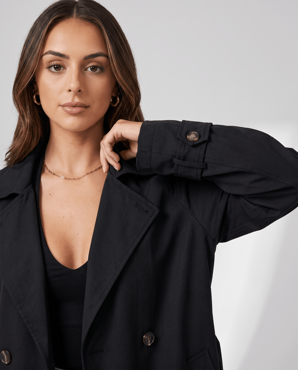 A woman wearing a black classic trench coat with a lapel collar and double-breasted button closure, adjusting the sleeve