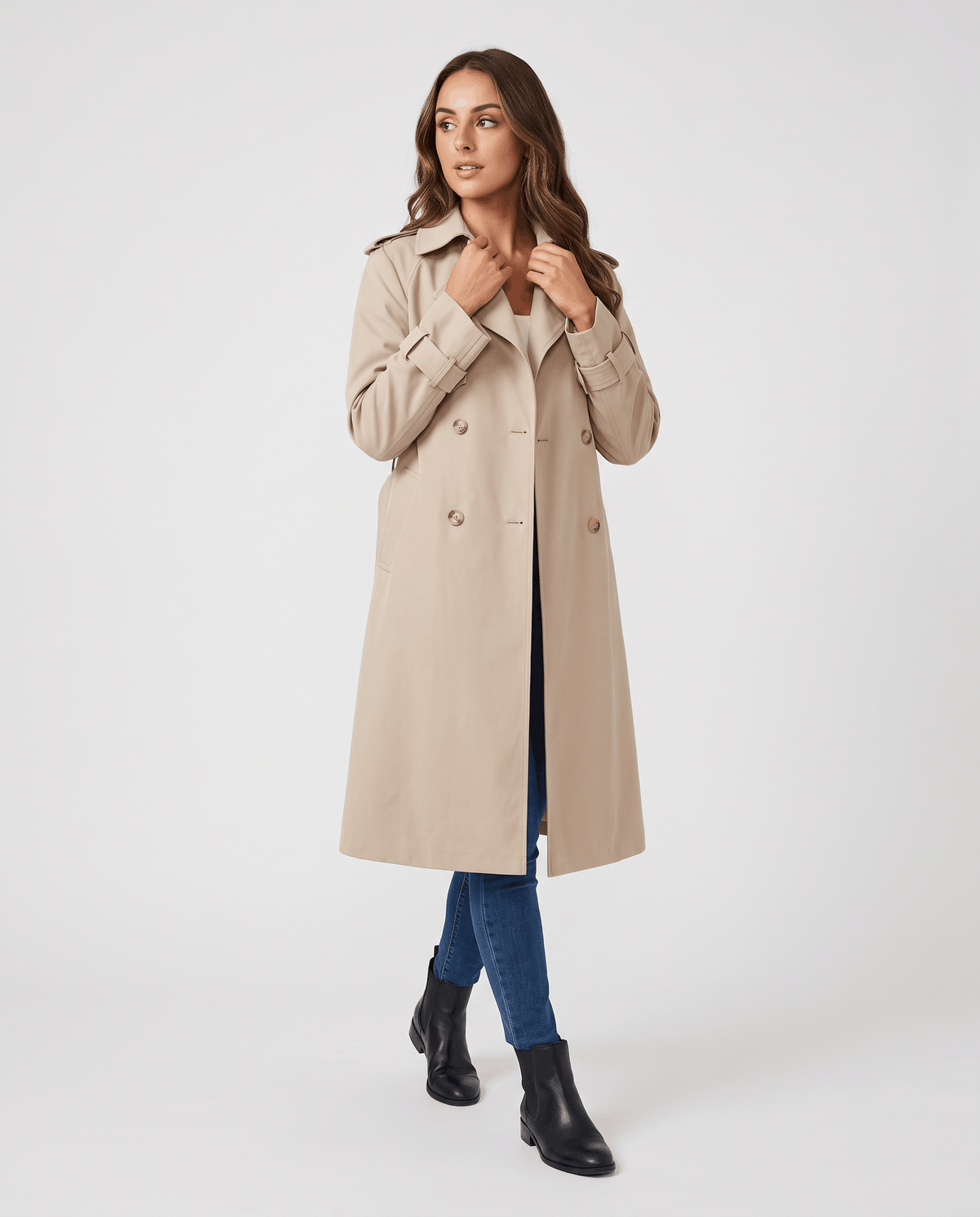 A woman wearing a beige classic trench coat with a lapel collar and double-breasted button closure, adjusting the collar