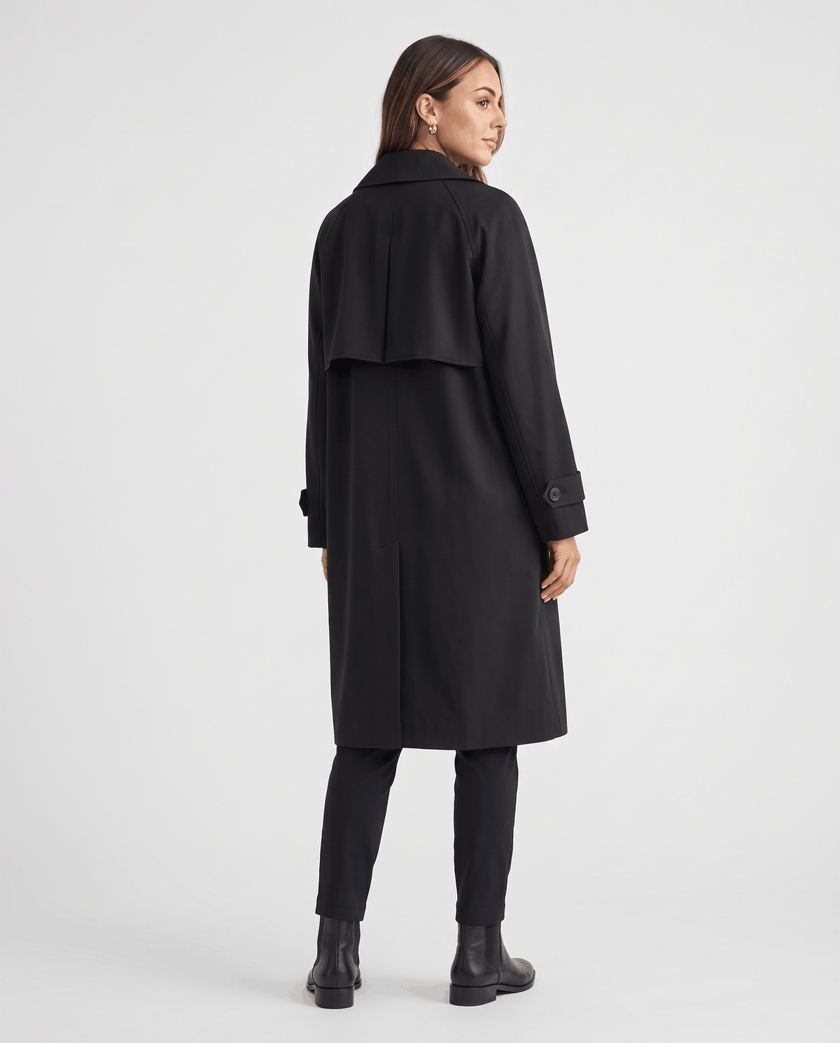 A woman wearing a black classic trench coat with a lapel collar and double-breasted button closure, shown from the back
