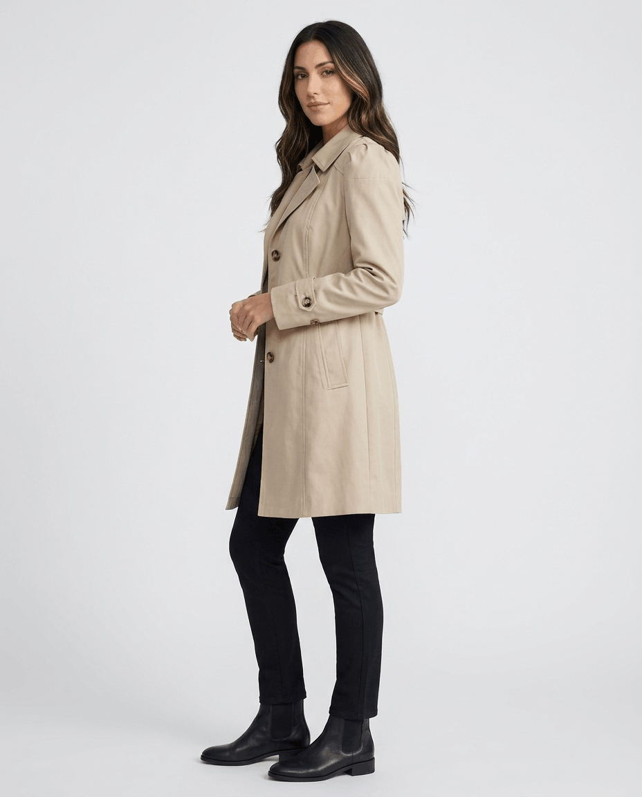A woman wearing a cream classic trench coat with a lapel collar, button front, and adjustable cuffs, side view