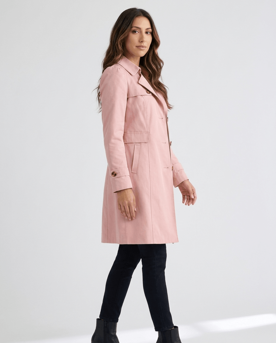 A woman wearing a pink classic trench coat with a lapel collar, button front, and adjustable cuffs, side view