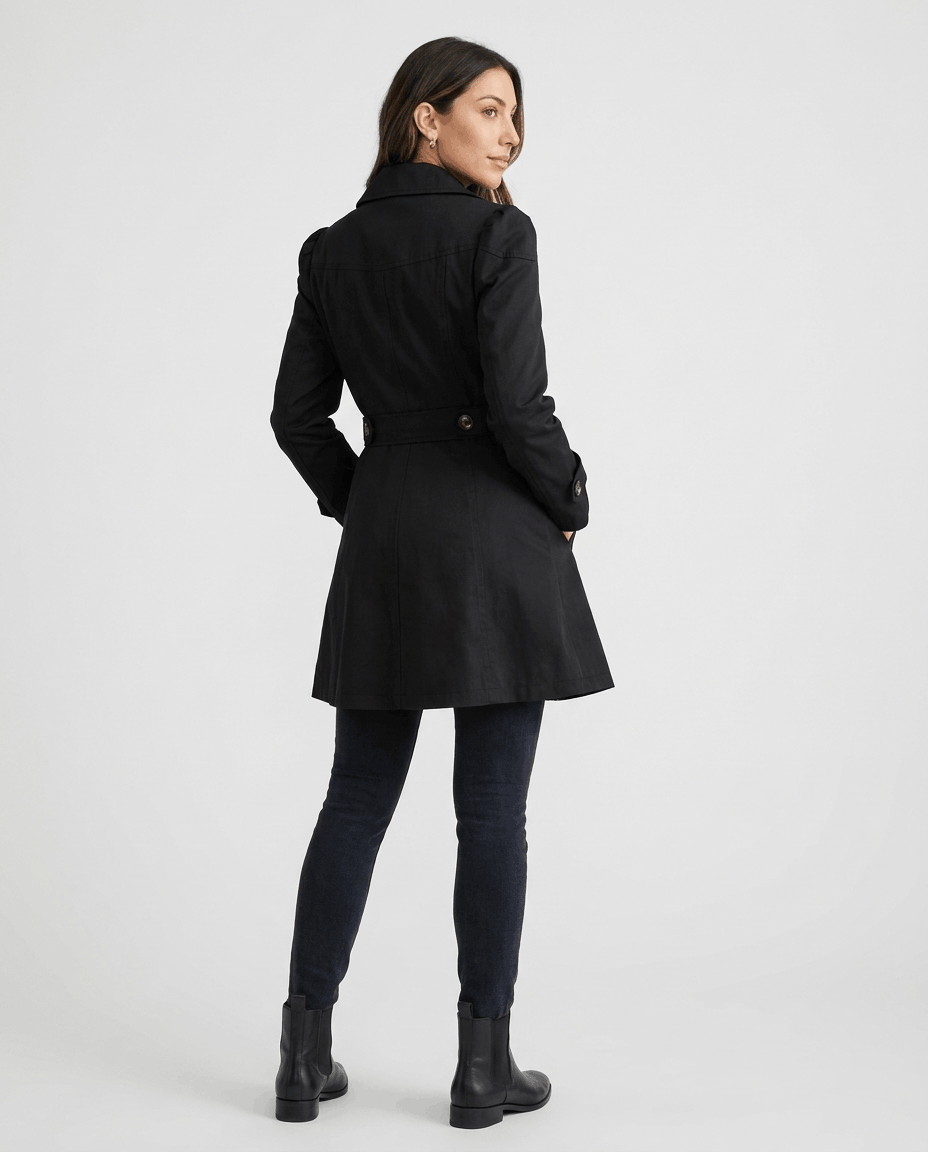 A woman wearing a black classic trench coat with a lapel collar, button front, and adjustable cuffs, back view