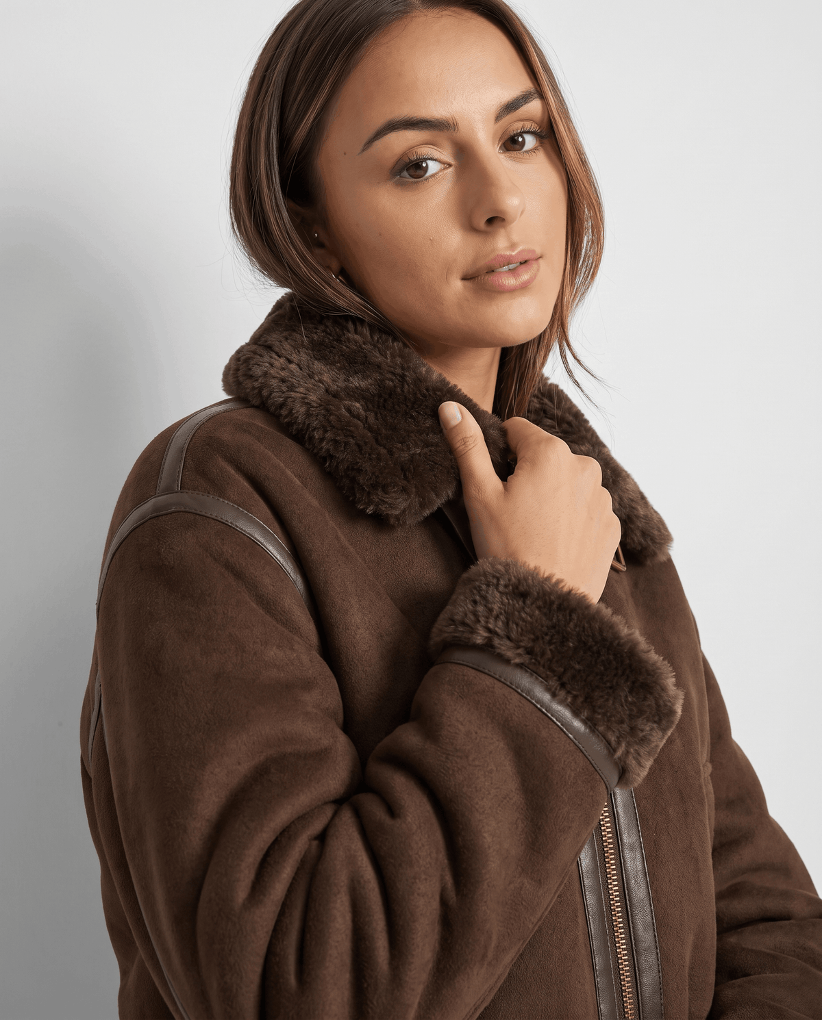 A woman wearing a brown cropped shearling bomber jacket with a plush collar and ribbed trim, adjusting the collar