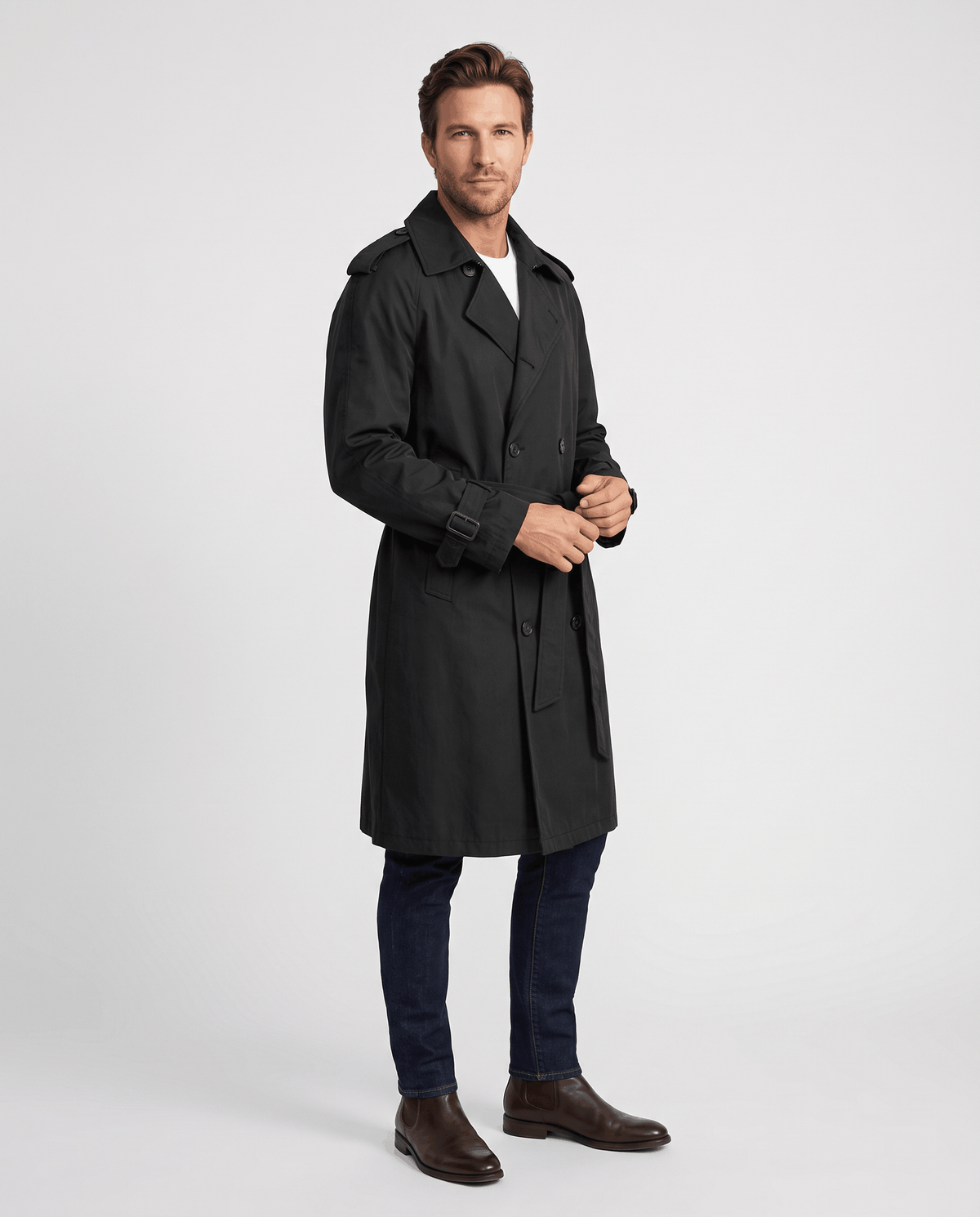 A man wearing a black double-breasted trench coat with a belted waist and adjustable cuffs, shown in side view