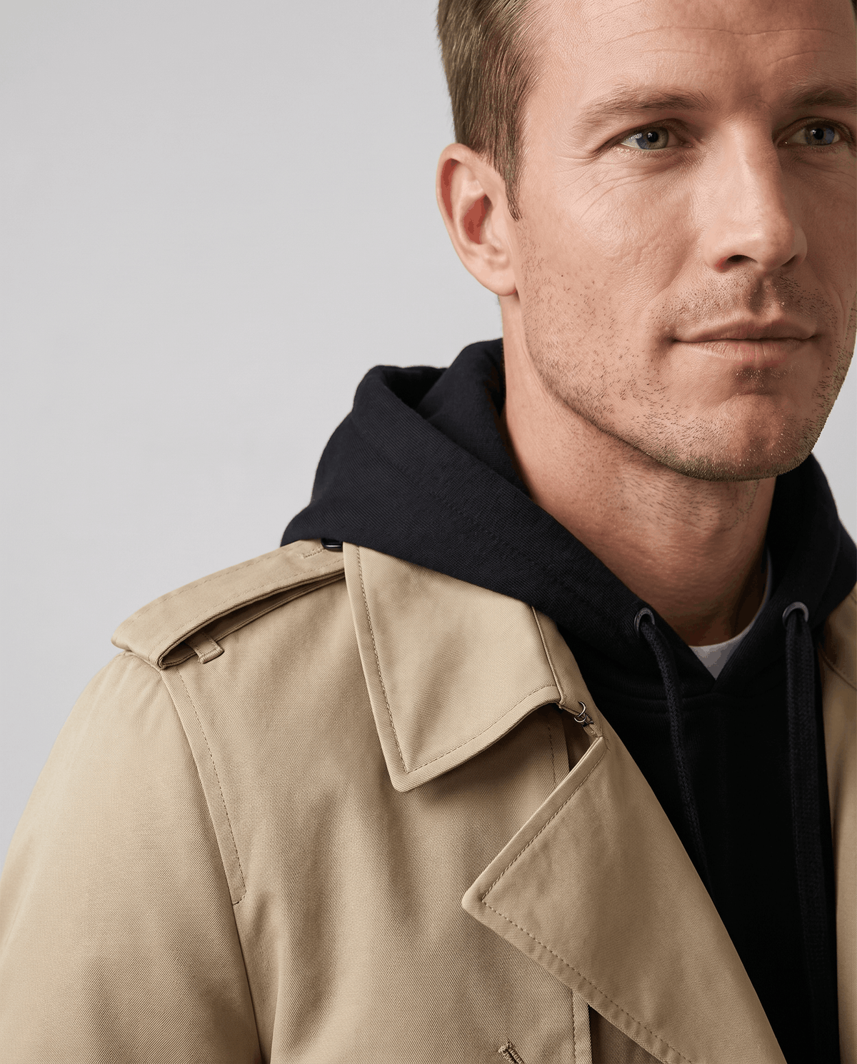 A man wearing a beige double-breasted trench coat with a notched lapel and shoulder epaulets, close-up view