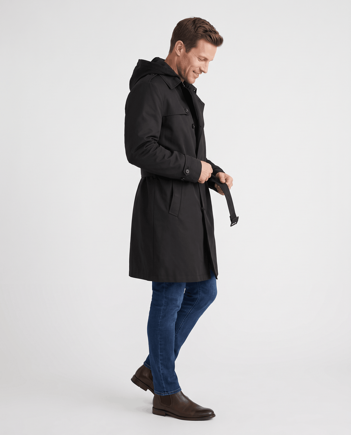 A man wearing a black double-breasted trench coat with a mid-length fit, notched lapel, and adjustable cuffs, side view