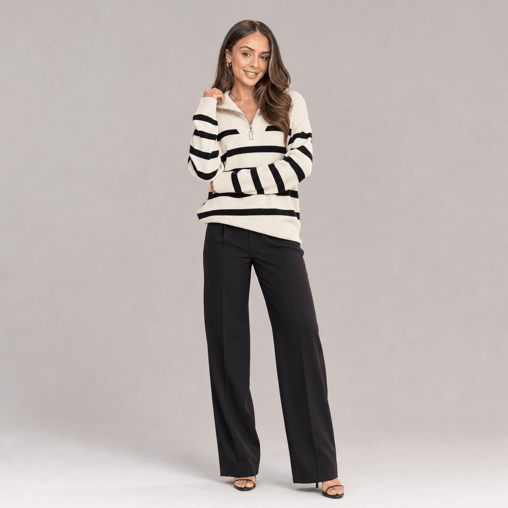 Full-body view of a female model wearing the cream and black striped quarter-zip sweater with black trousers.