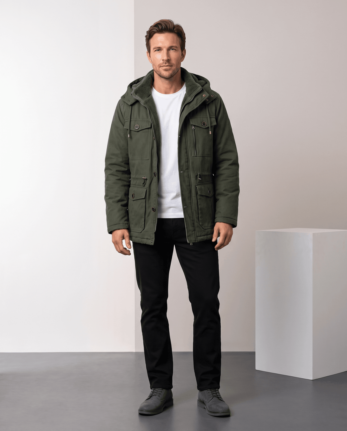 A man wearing a dark green fleece-lined parka with a detachable hood, insulated design, and multiple utility pockets