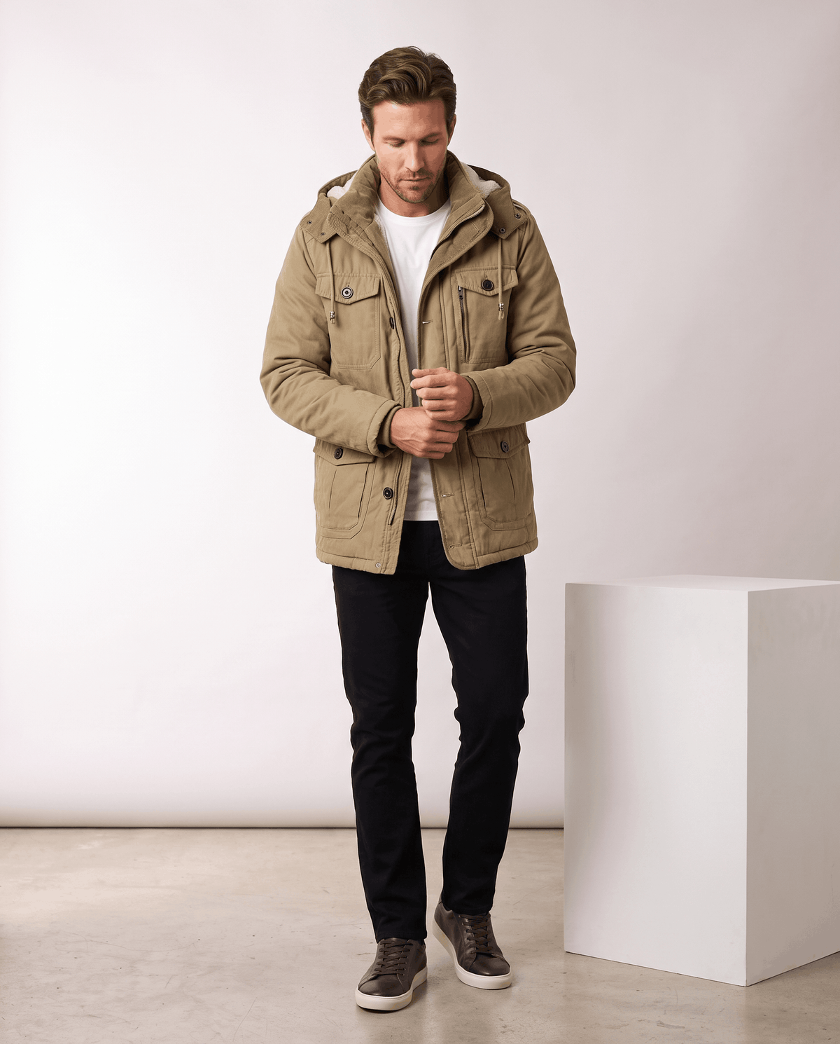 A man wearing a beige fleece-lined parka with a detachable hood and insulated design, standing indoors