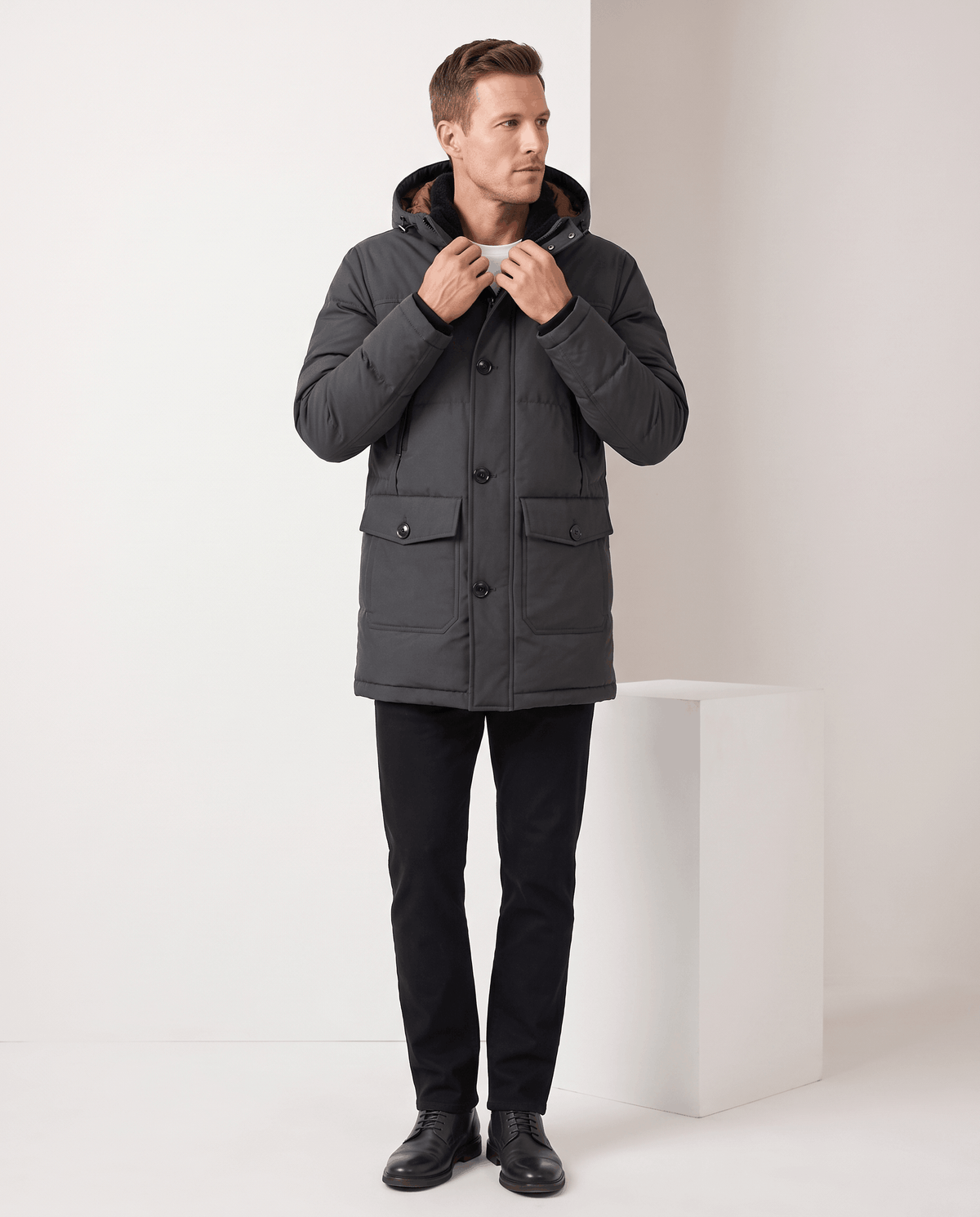 A man wearing a dark gray insulated winter parka with a hood, full-zip front, and multiple utility pockets, standing indoors