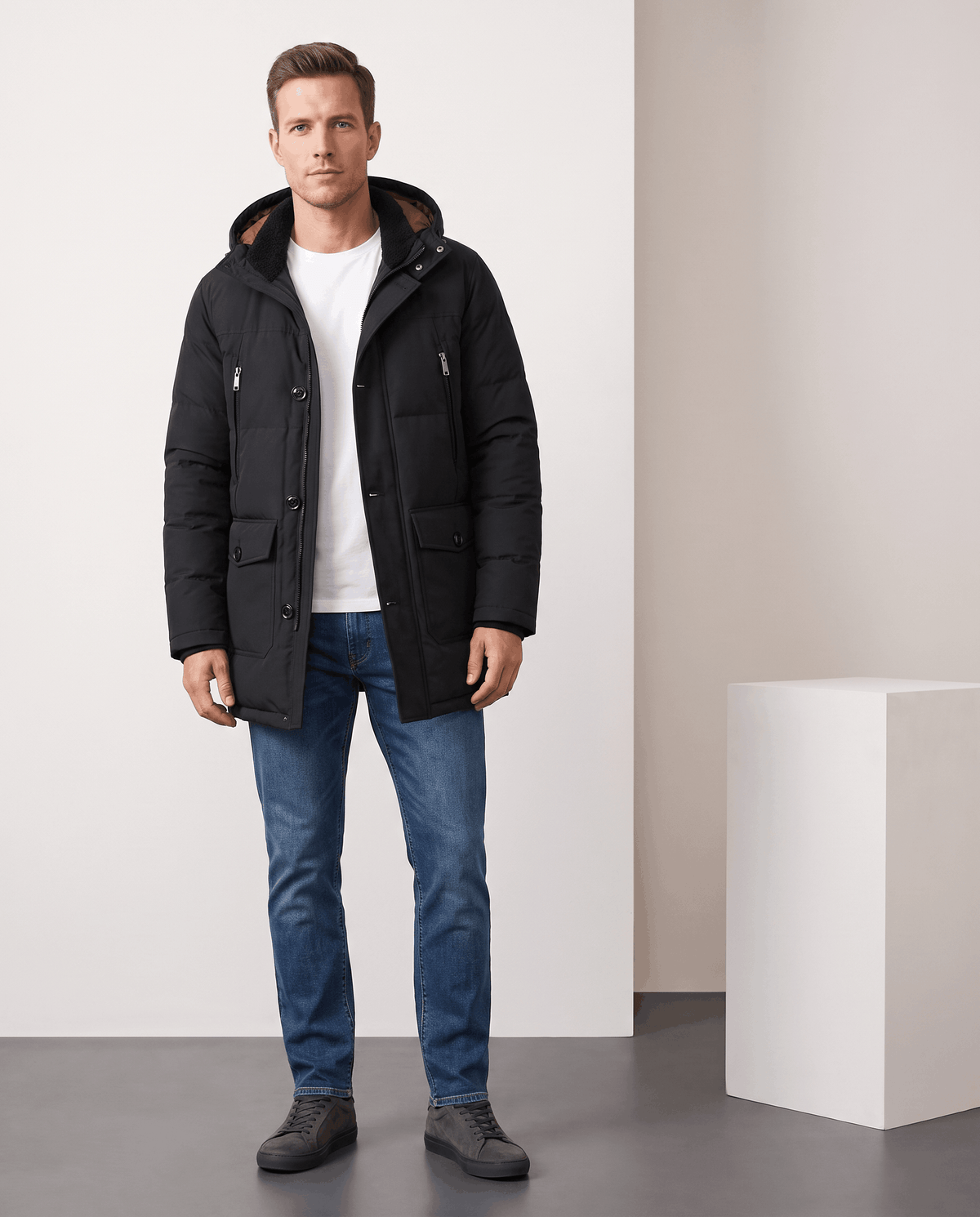 A man wearing a black insulated winter parka with a hood and multiple utility pockets, standing indoors