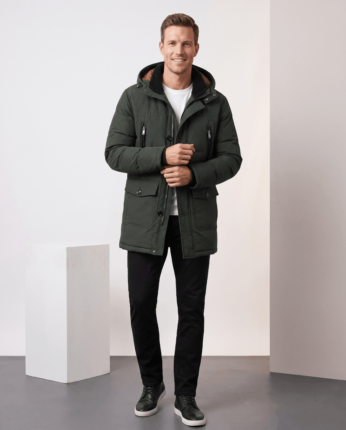 A man wearing a dark green insulated winter parka with a hood and multiple utility pockets, standing indoors