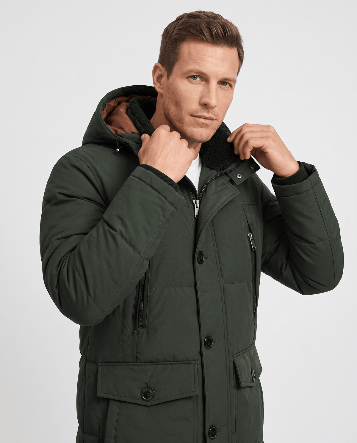 A man wearing a dark green insulated winter parka with a hood and multiple utility pockets, adjusting the hood