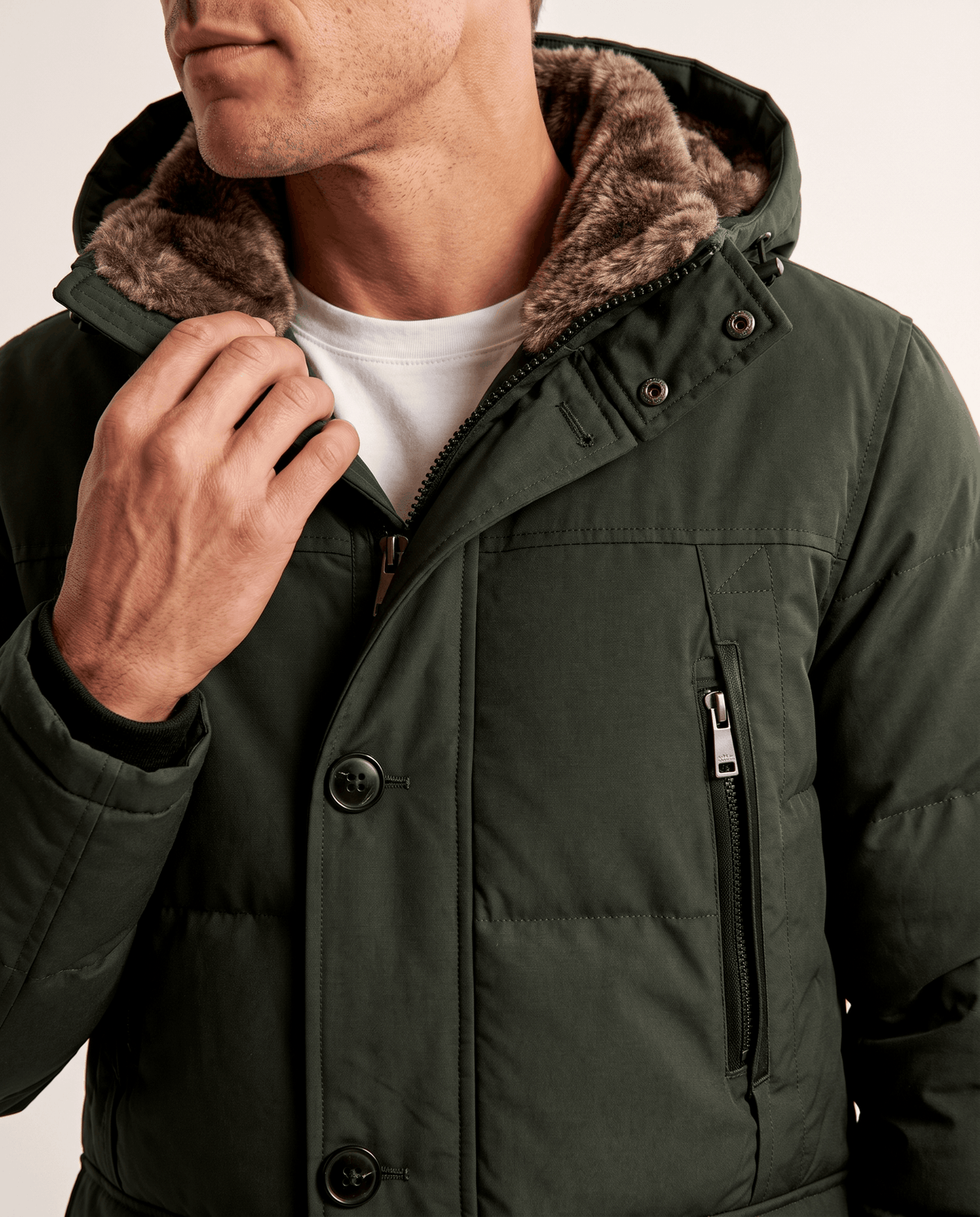 Close-up of a man wearing a dark green insulated winter parka, showing the hood, collar, and front utility pocket