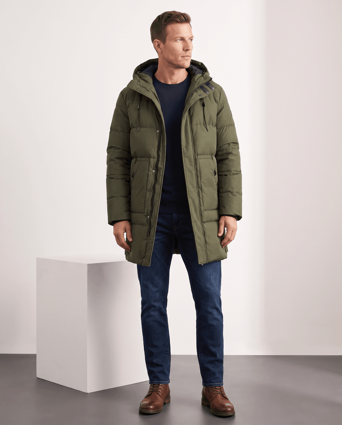 A man wearing an olive green insulated winter parka with a hood and padded longline design, standing indoors