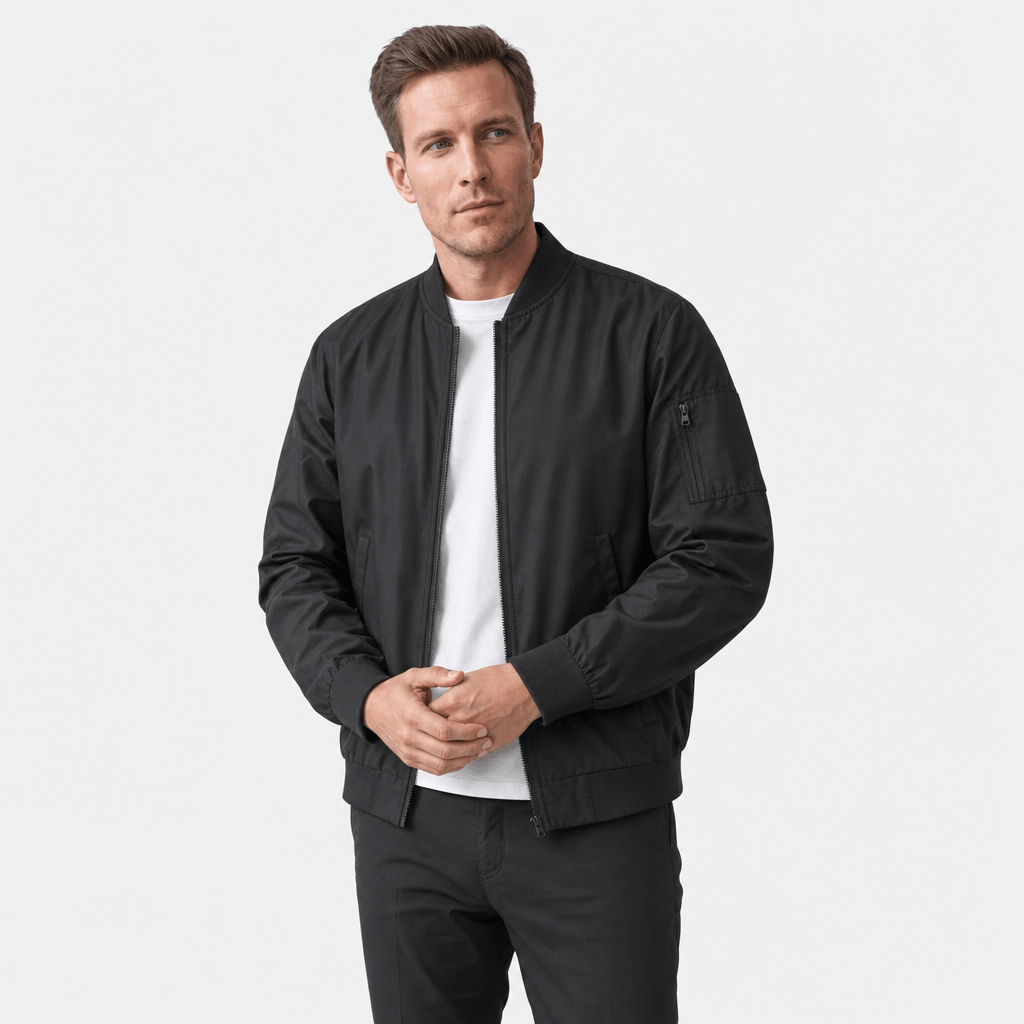 Man wearing a black bomber jacket over a white T-shirt and black trousers, standing against a light studio background.