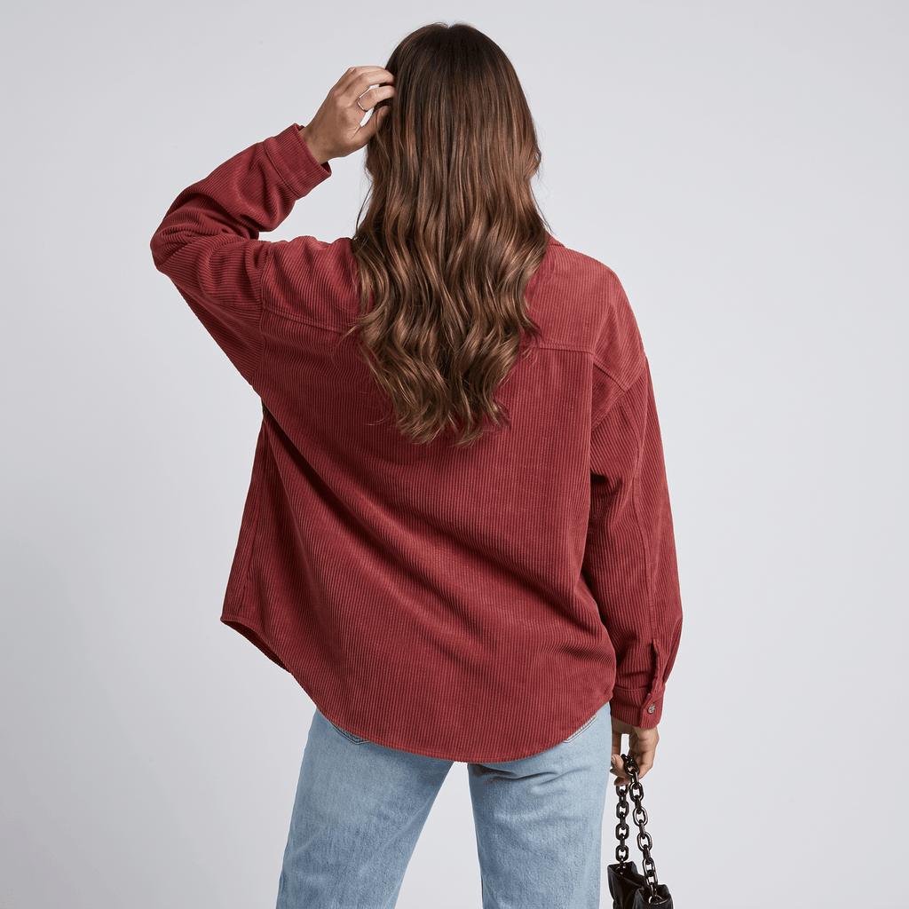 Rust-toned oversized corduroy button-down shirt with a drop-shoulder fit and ribbed texture, worn with light-wash jeans.