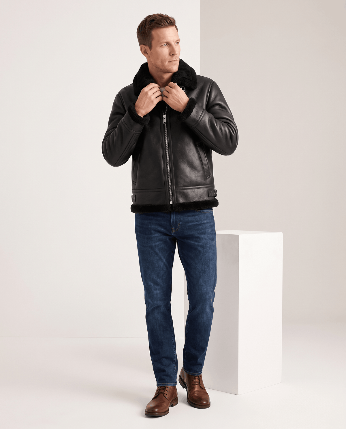 A man wearing a black leather shearling aviator jacket with an asymmetrical zip and buckle strap, adjusting the collar