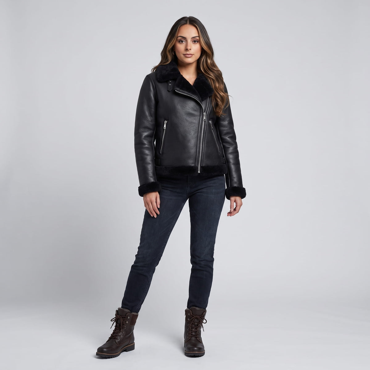 A woman wearing a black leather shearling biker jacket with an asymmetrical zip and plush trim, standing indoors