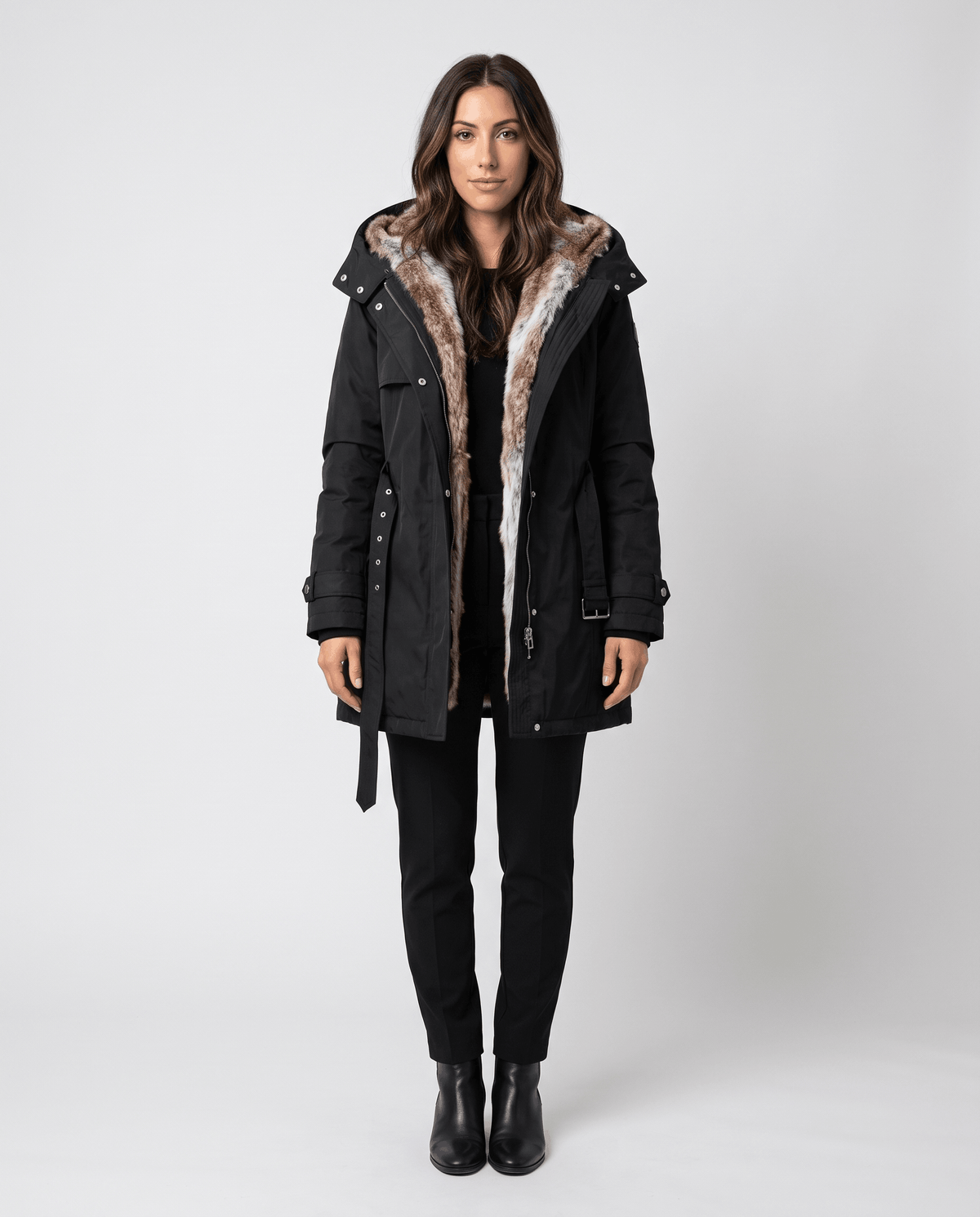A woman wearing a black long parka coat with fur lining, a hood, and an adjustable waist, front view