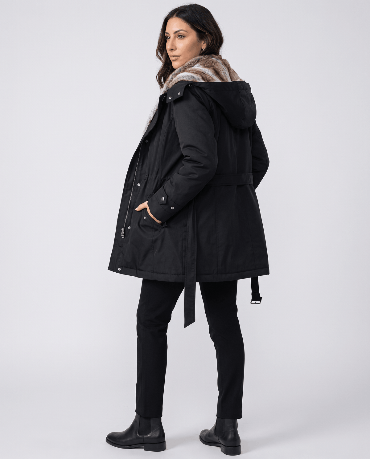A woman wearing a black long parka coat with fur lining, a hood, and an adjustable waist, side view