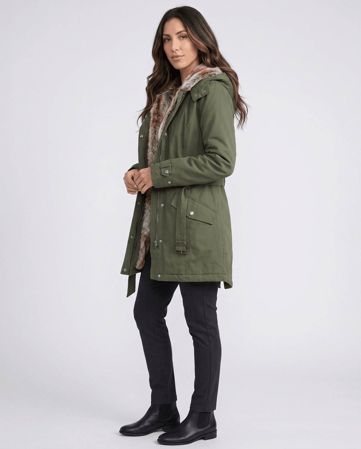 A woman wearing an olive green long parka coat with fur lining, a hood, and an adjustable waist, full-body view
