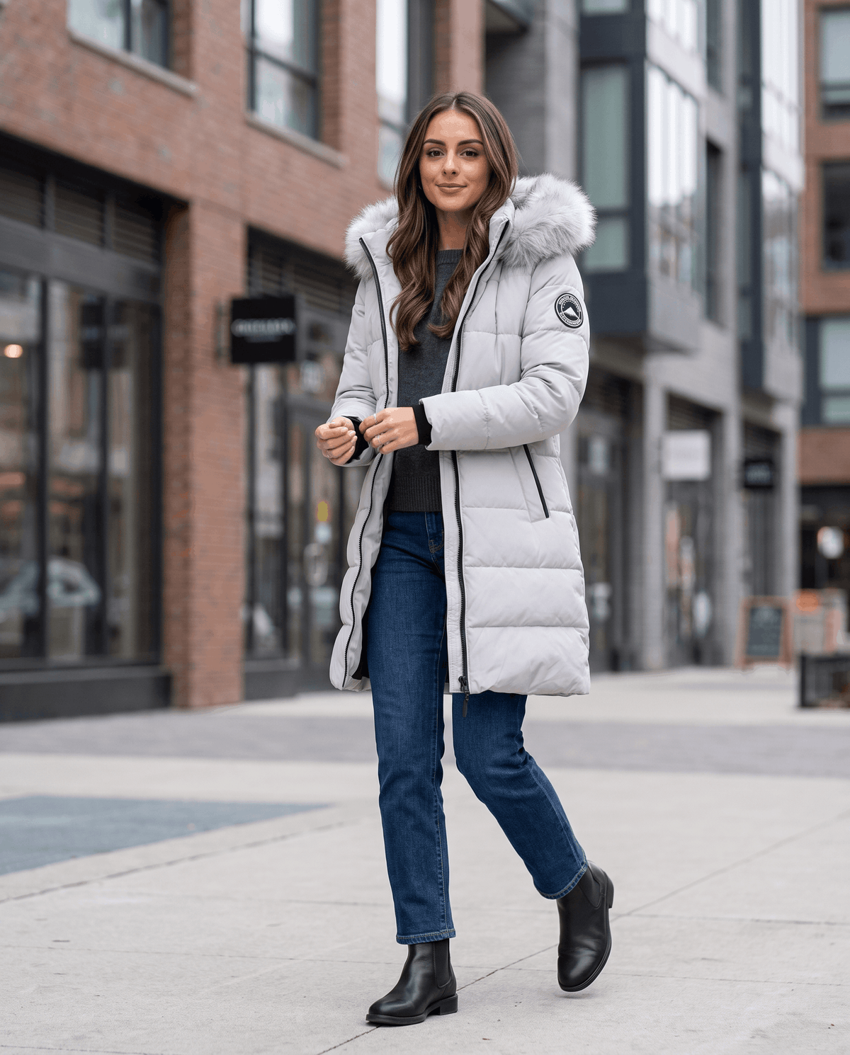 A woman wearing a light gray long insulated parka jacket with a fur-trimmed hood and full-zip front, walking on a city street