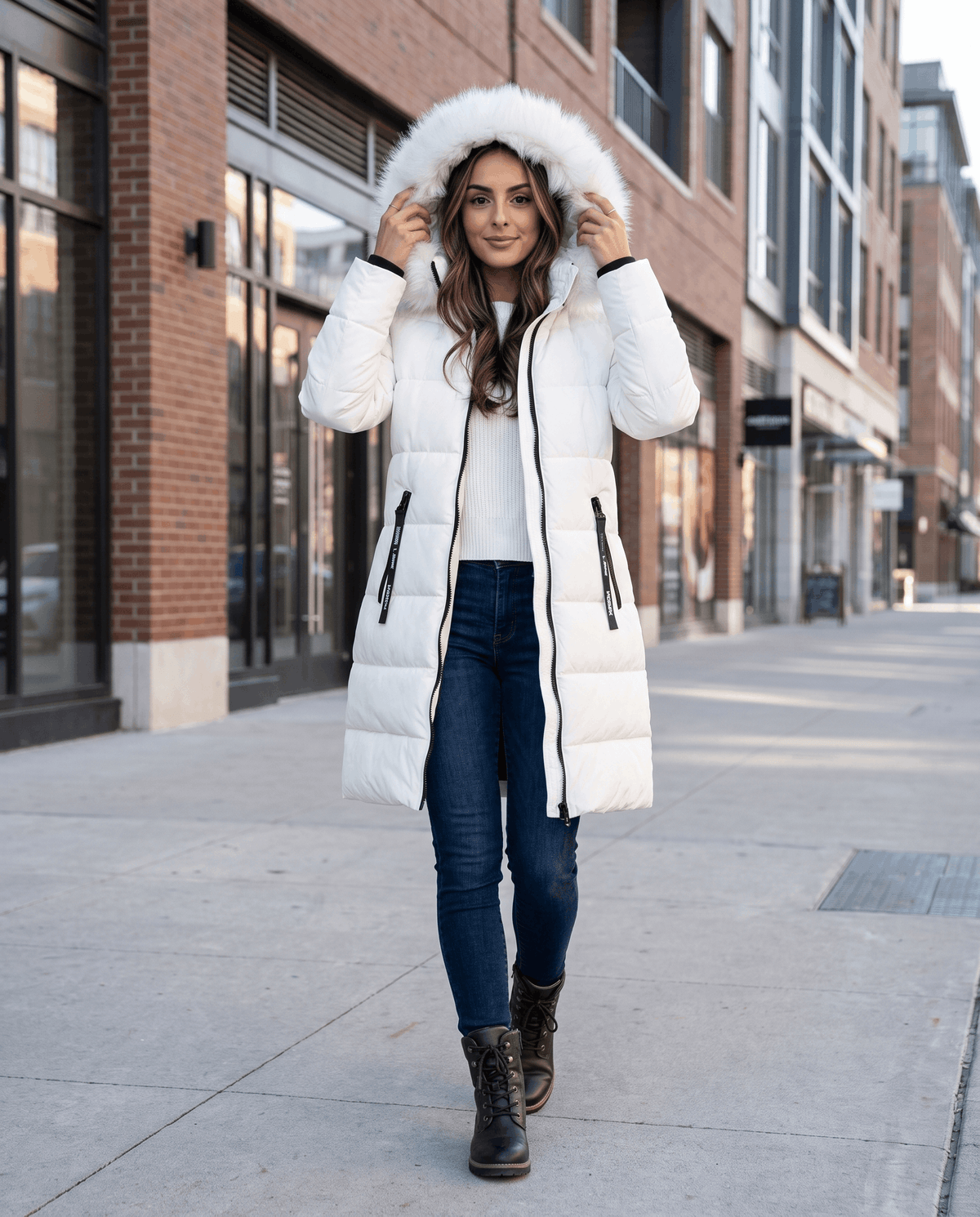 A woman wearing a white long insulated parka jacket with a fur-trimmed hood and full-zip front, walking on a city street