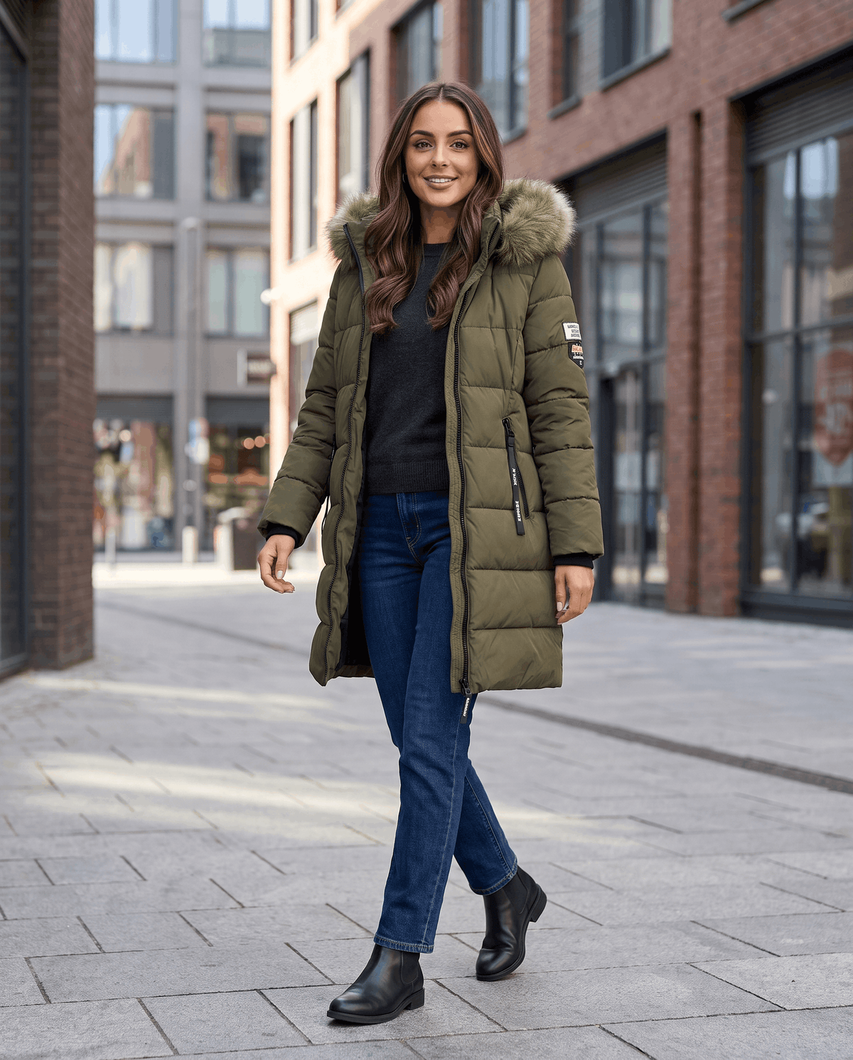 A woman wearing a long olive green insulated parka jacket with a hood and full-zip front, walking on a city street